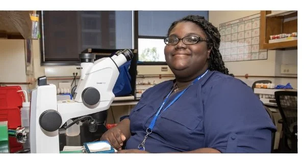 A woman in blue scrubs and glasses sitting at a lab bench with a microscope in a laboratory setting.
