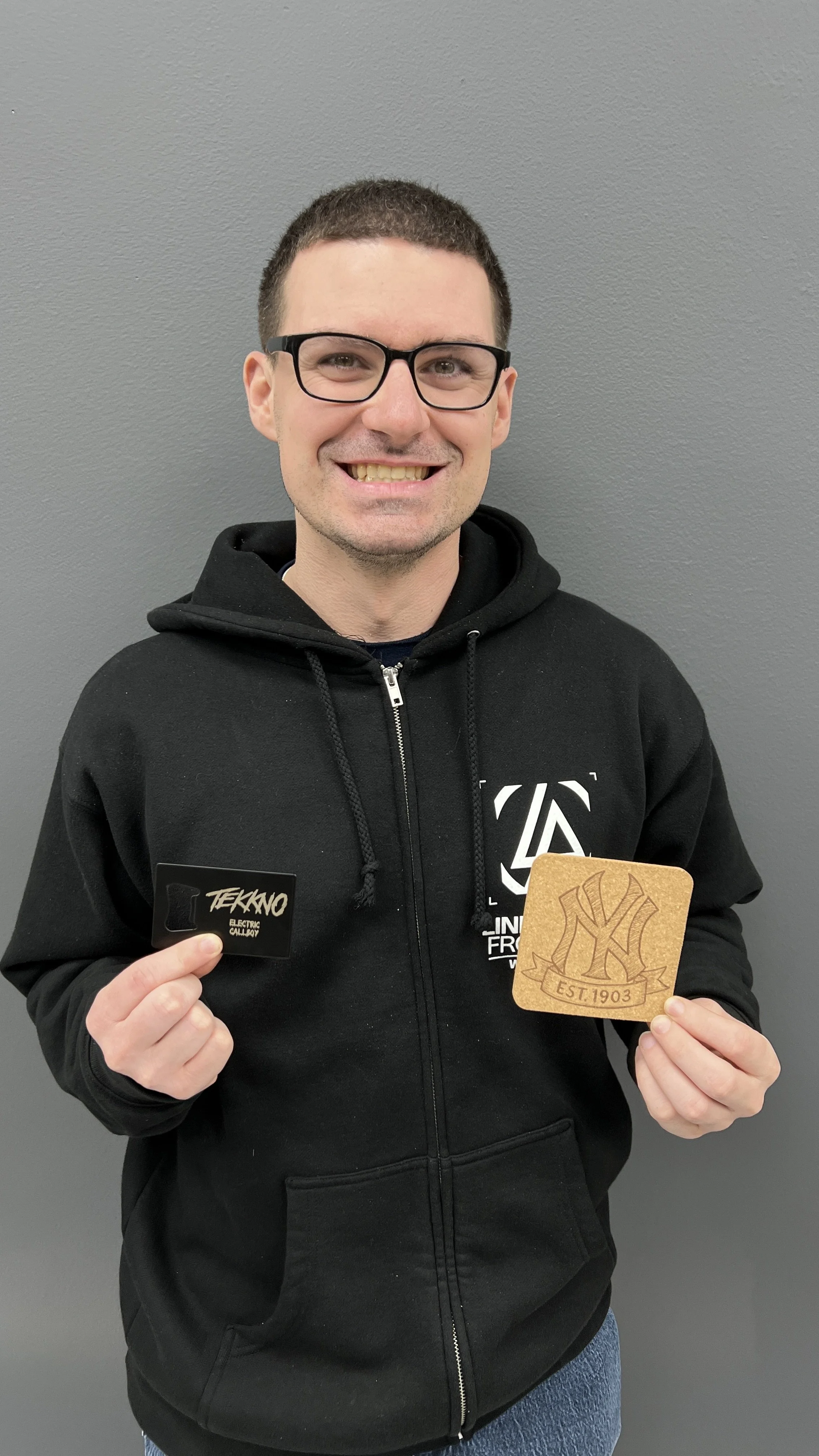 A smiling man with glasses, wearing a black hoodie, holding a black card in one hand and a wooden coaster with a New York Yankees logo in the other, standing against a gray wall.
