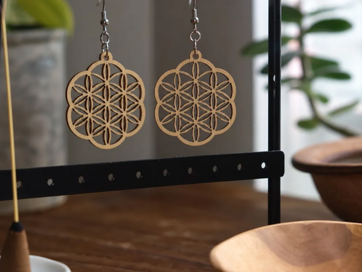 Pair of wooden earrings with geometric floral pattern hanging on a metal display stand in a room with wooden and green elements.