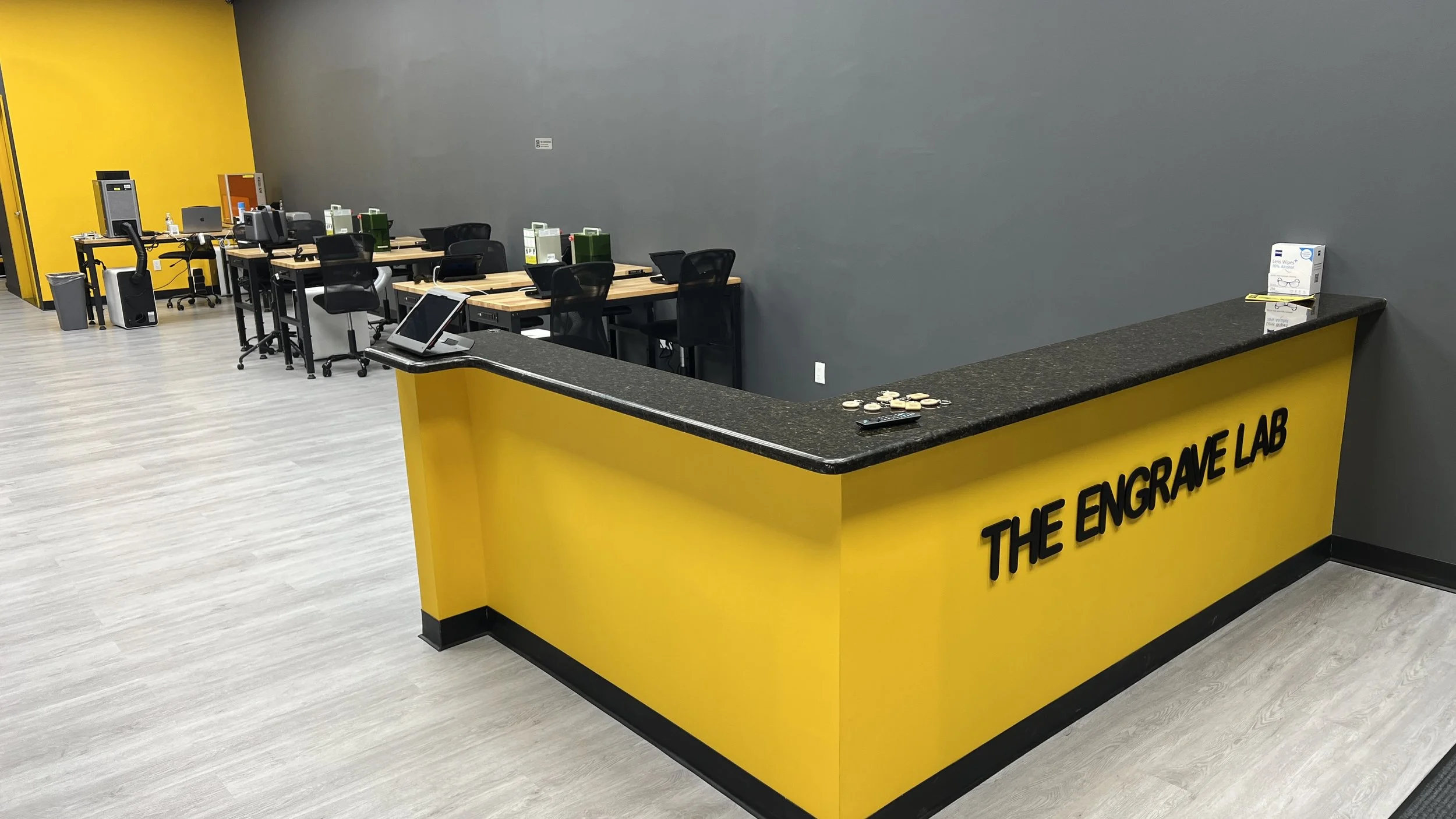 Reception desk with yellow front panel labeled 'The Engrave Lab' in black text, with some coins and a remote on top. Behind the desk, there are multiple workstations with chairs, computers, and office supplies, set against a gray wall and a yellow accent wall.