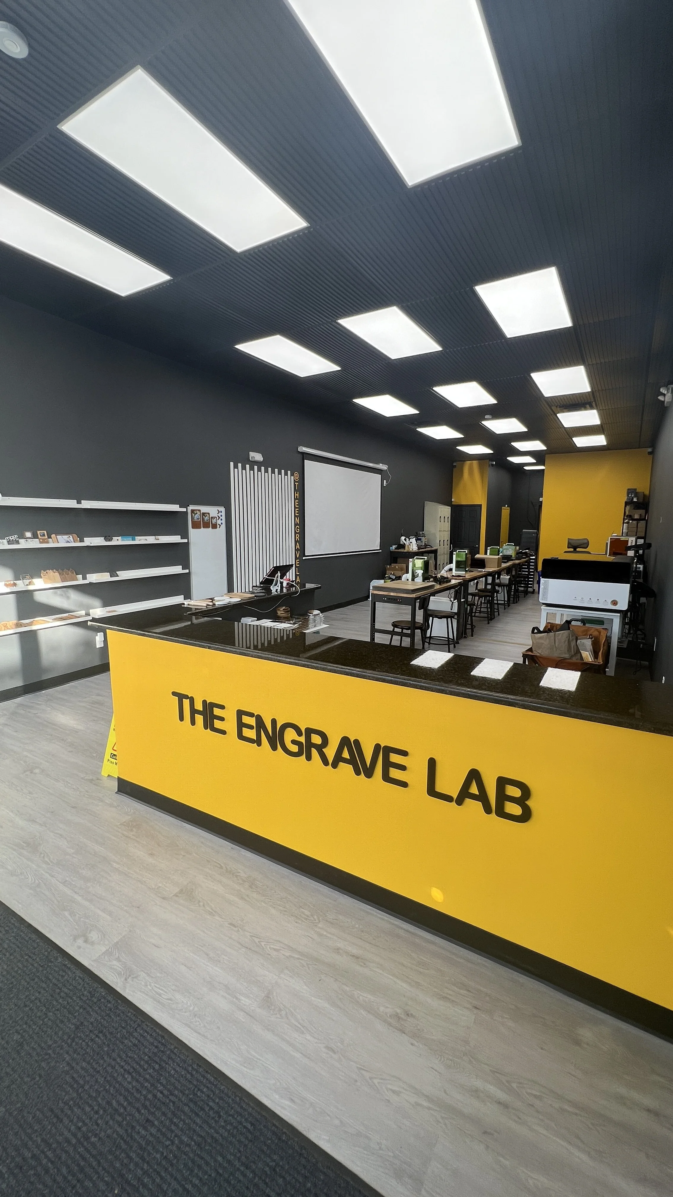 Interior view of an engraving lab with a yellow counter reading "The Engrave Lab," shelves with samples, a white projection screen, multiple workstations with computers, and industrial equipment against a black and yellow wall.