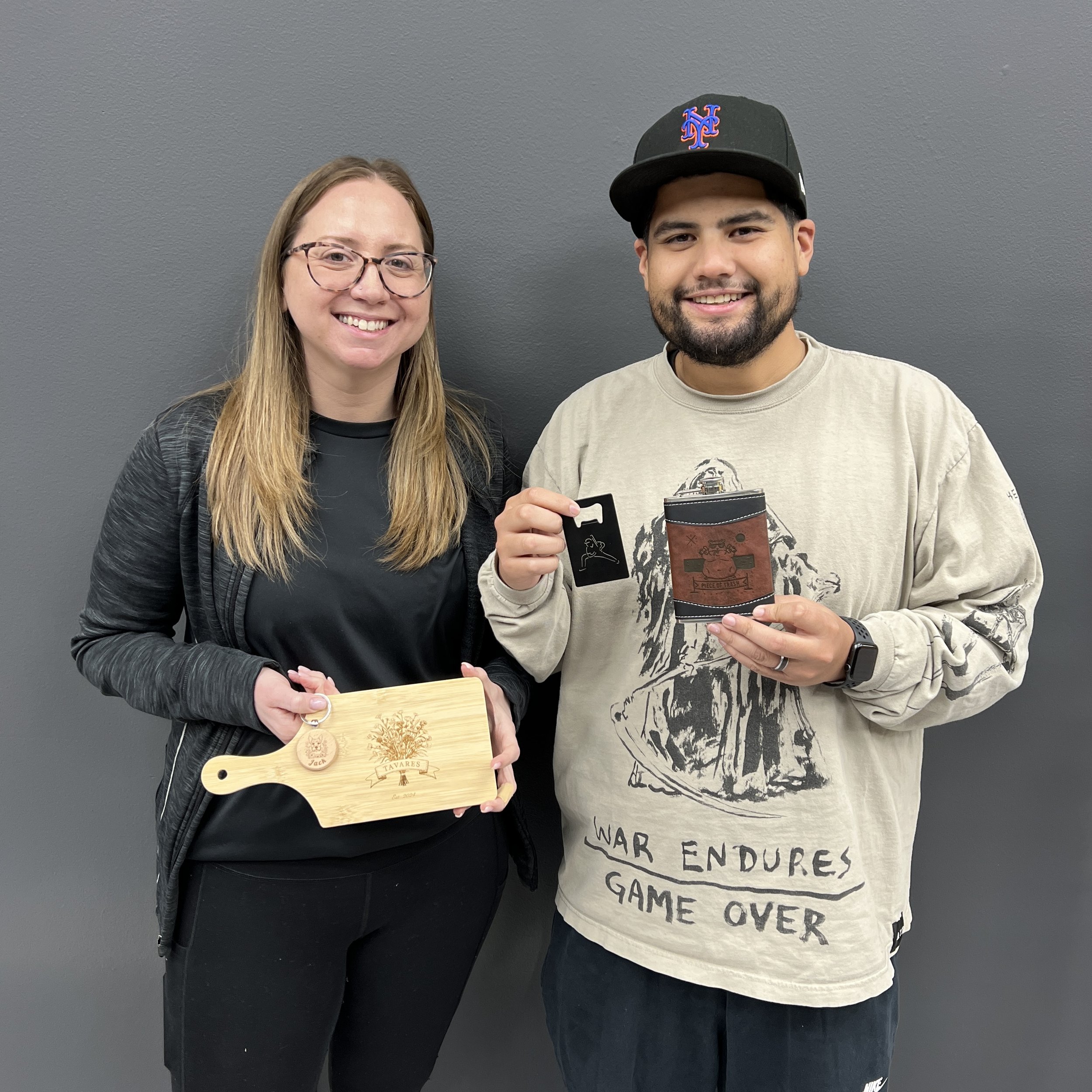 A woman and a man standing against a gray wall, smiling, holding wooden and leather items, with the woman holding a wooden cutting board and the man holding a leather wallet and keychain.