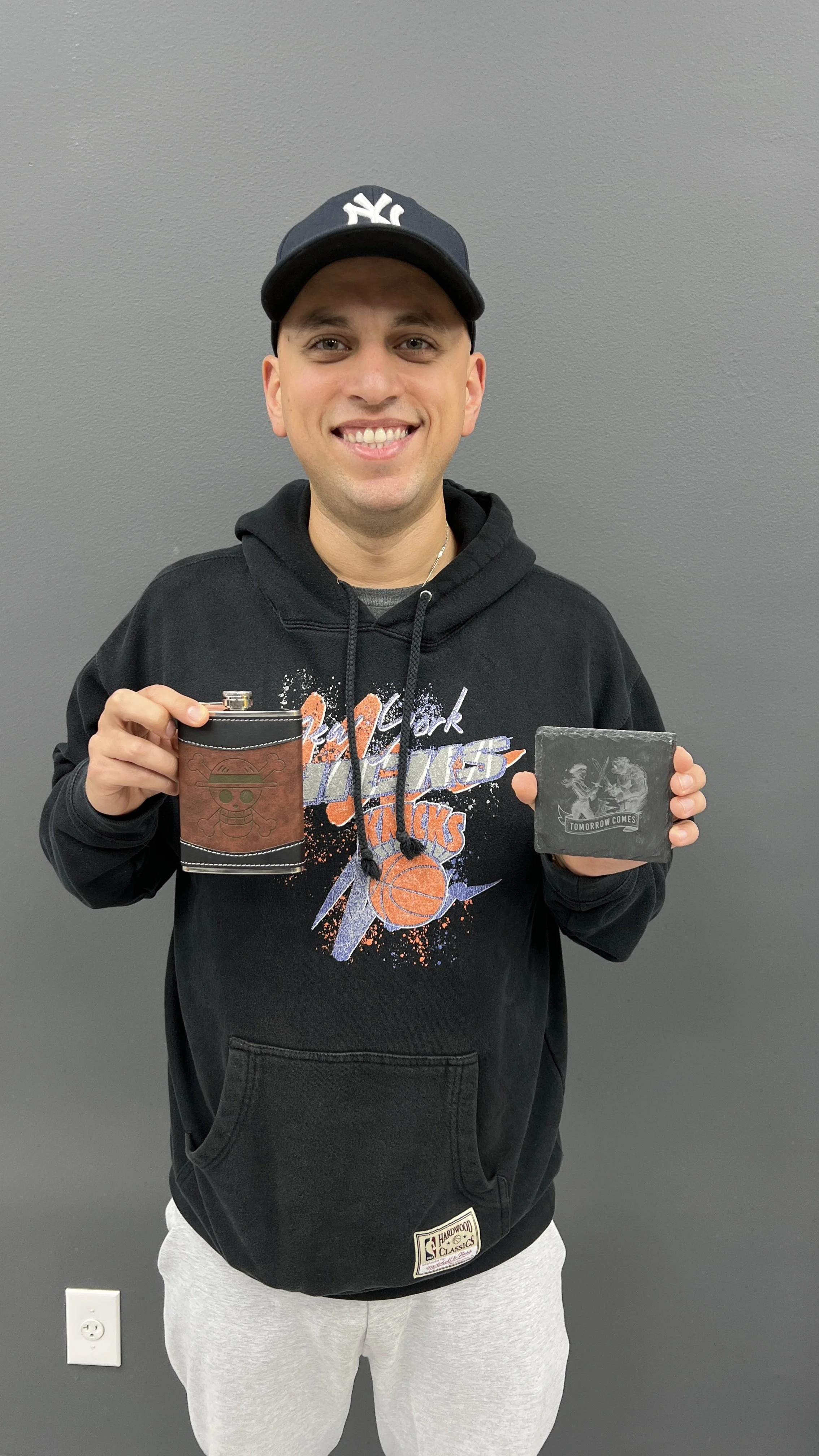 A young man wearing a New York Knicks hoodie and a New York Yankees cap, smiling and holding a flask with a skull and crossbones design in one hand and a black box with a skeleton graphic and the text 'Tomorrow Comes' in the other.