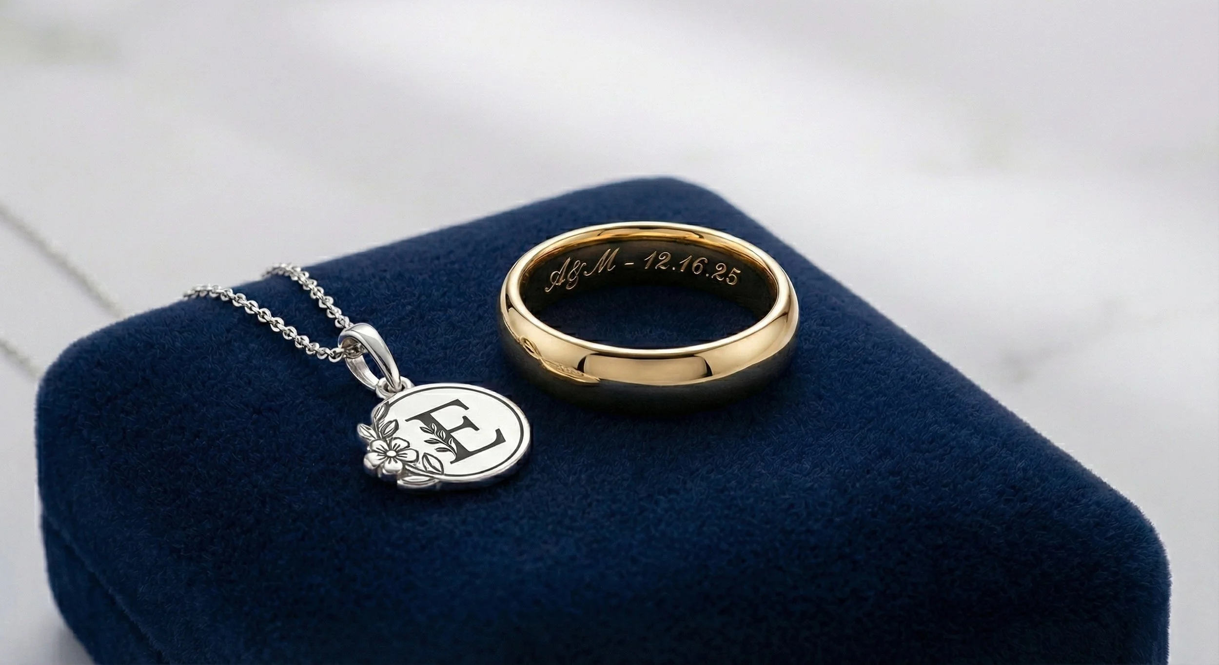 A silver necklace with a circular pendant and a gold wedding band engraved with initials and a date, displayed on a dark blue cushion.