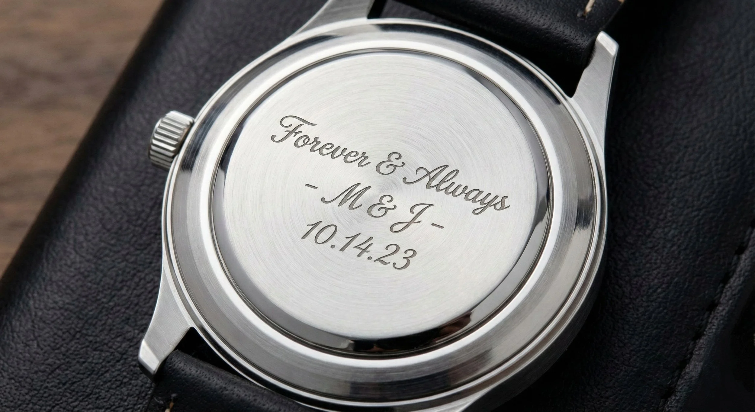 The back of a wristwatch with engraved text reading, "Forever & Always - M & J - 10.14.23", on a black strap, placed on black textured surface.