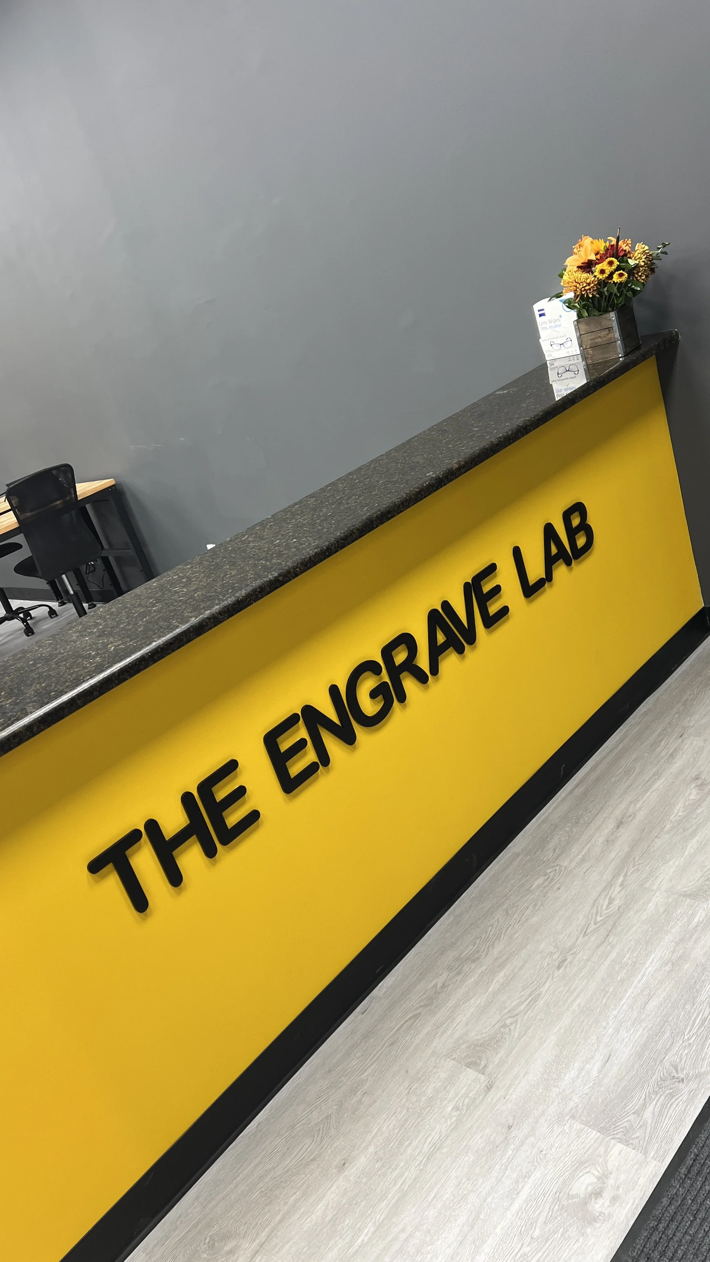 The reception desk of The Engrave Lab with a yellow front panel, some flowers, and a pair of glasses on a small card holder. There are chairs and tables in the background.
