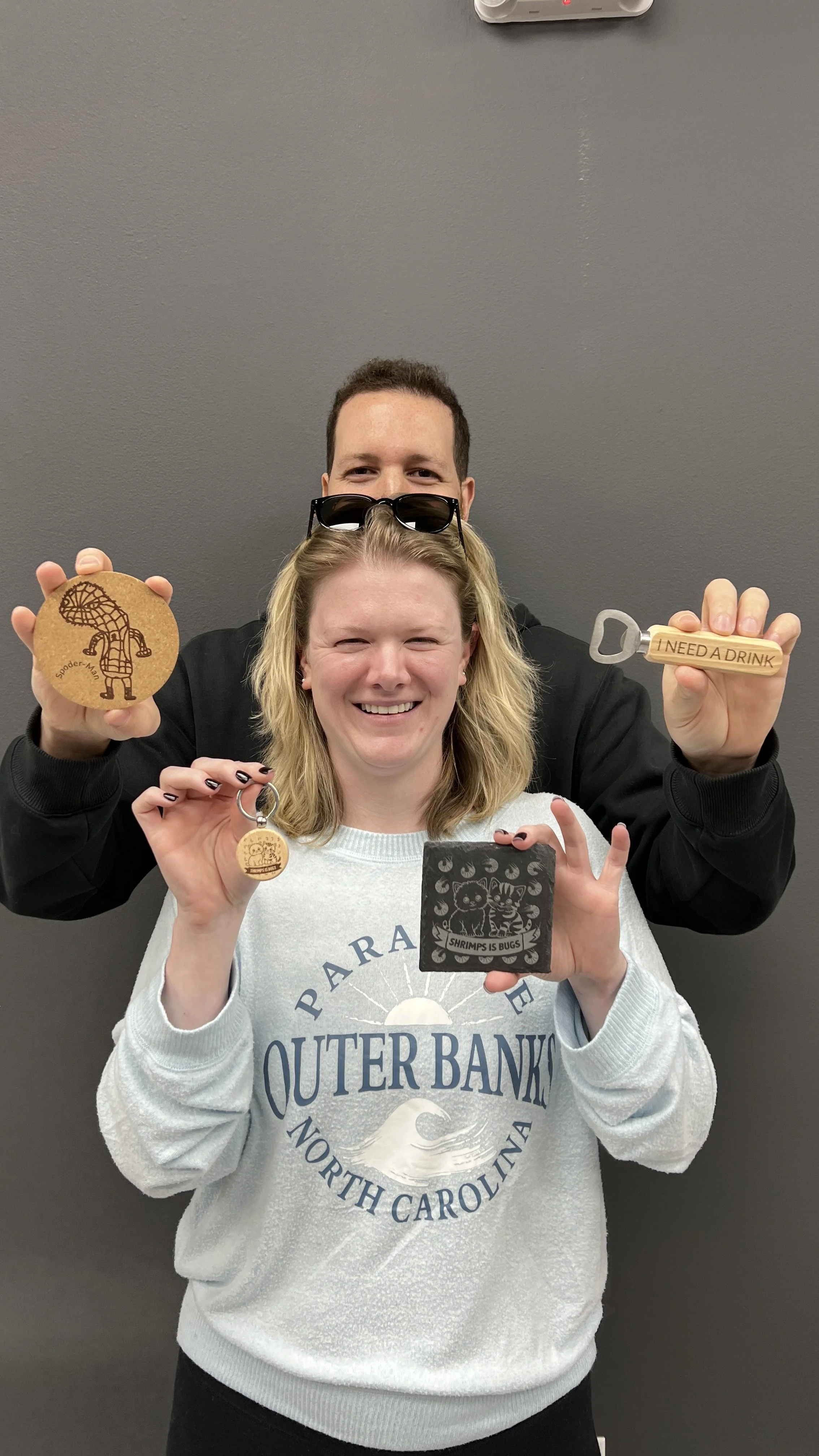 Two smiling people, a man and a woman, holding various wooden collectibles. The woman, in front, is holding a small round wooden keychain with a design, and a square coaster with a tiger and the text 'SHRIMPIS IS BUGS.' The man behind her is holding 