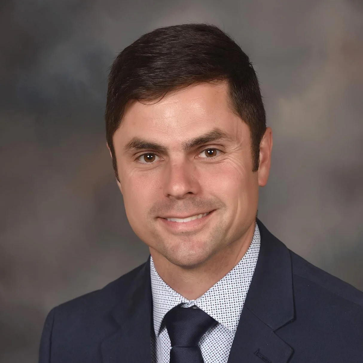 Professional headshot of a man with short dark brown hair, wearing a dark suit, white shirt with a dotted pattern, and a dark tie, against a blurred gray background.