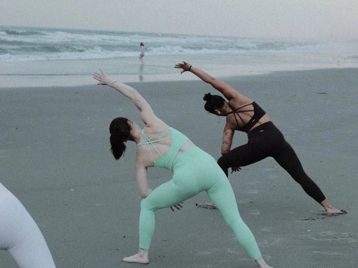 Two women practicing yoga on the beach, in side stretches, with ocean waves in the background.
