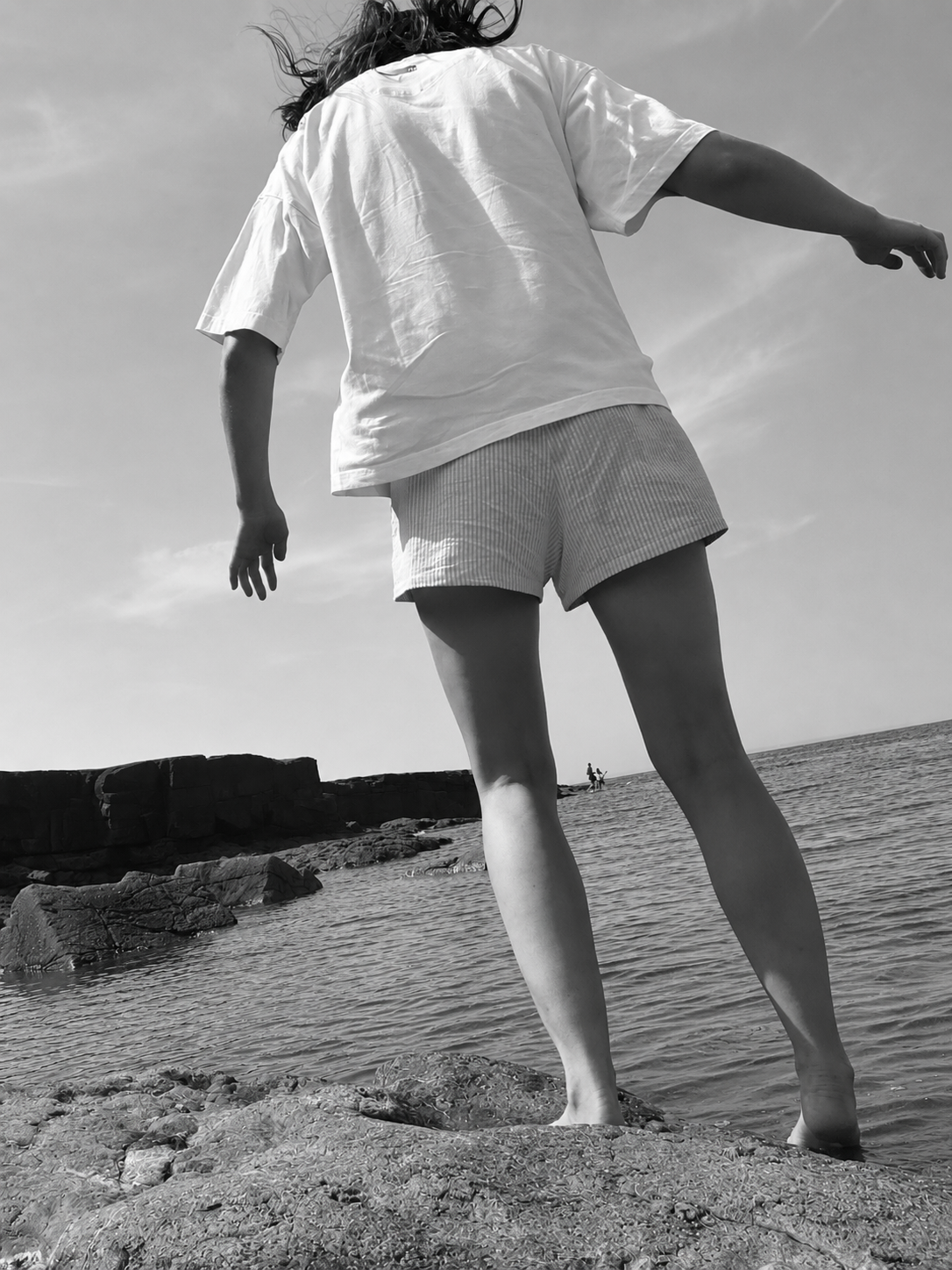 A woman in a white t-shirt and striped shorts standing on rocks at the beach, facing the water, with the sky visible in the background.