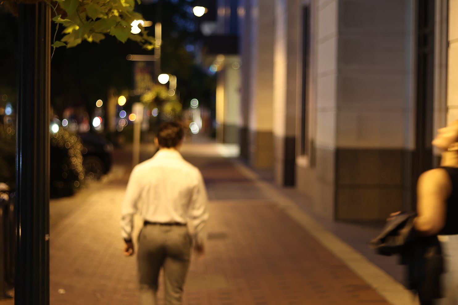 A person walking alone on a city sidewalk at night, blurry streetlights and buildings in the background.