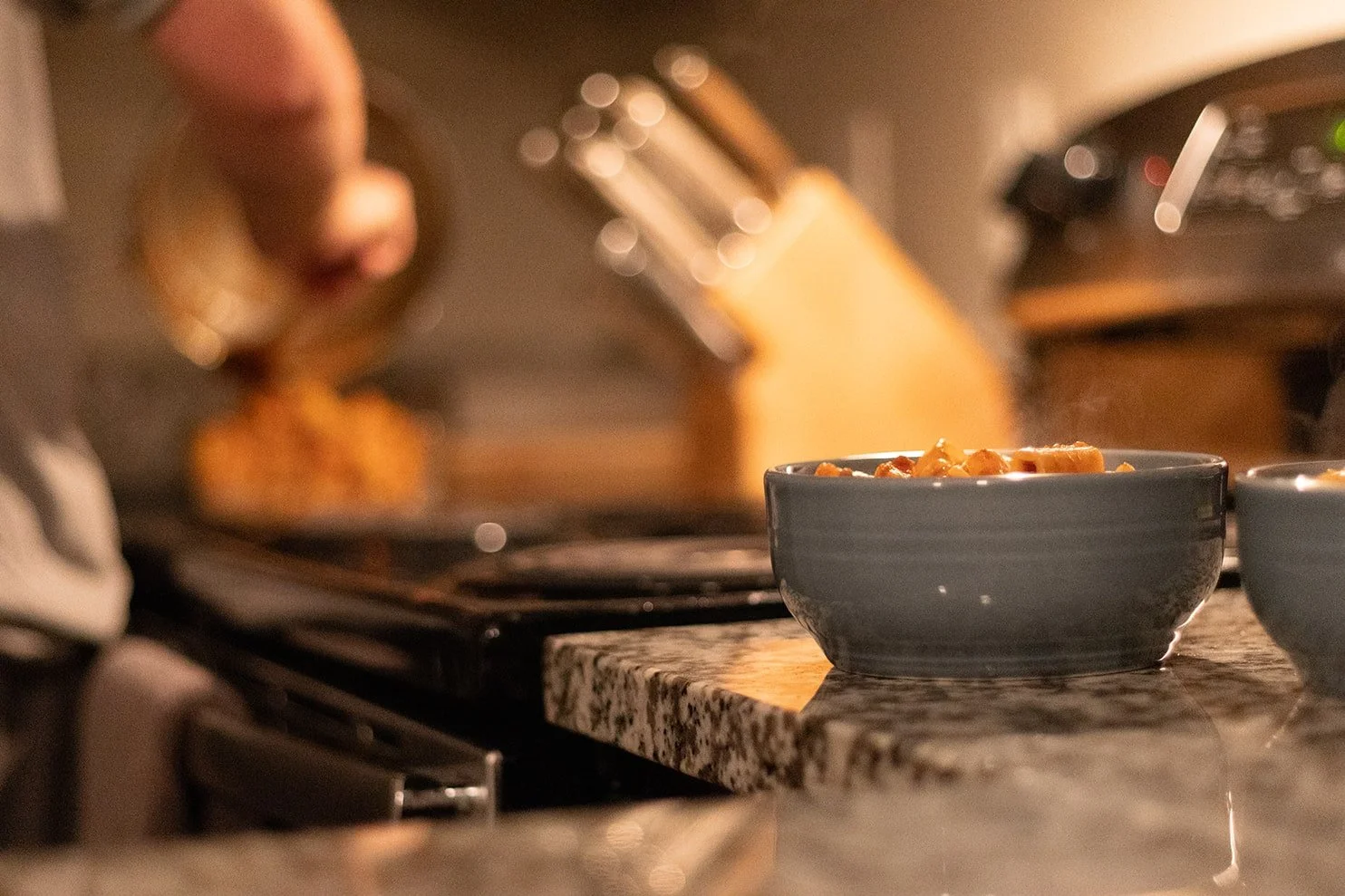 Close-up of a kitchen counter with two bowls of food in the foreground and a person cooking in the background, with a stove and oven.