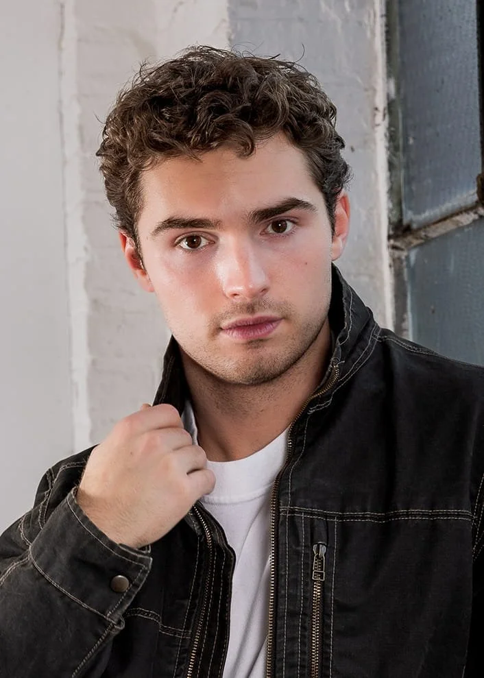 A young man with curly brown hair and light skin, wearing a black jacket over a white shirt, is looking at the camera and holding the collar of his jacket, standing in front of a white and metallic background.