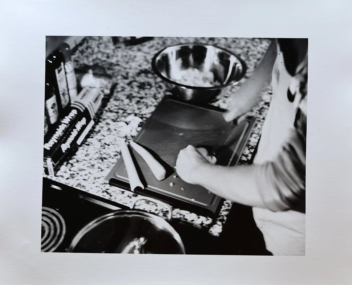 Black and white photo of a person using a digital scale on a kitchen countertop with various kitchen items including a bowl, a box, a spoon, and a frying pan.