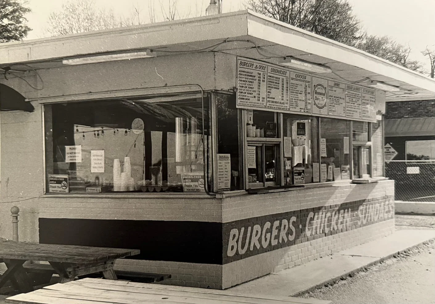 A vintage fast food restaurant serving burgers, grilled sandwiches, and chicken, with a large sign displaying the menu above the counter and a window for ordering.