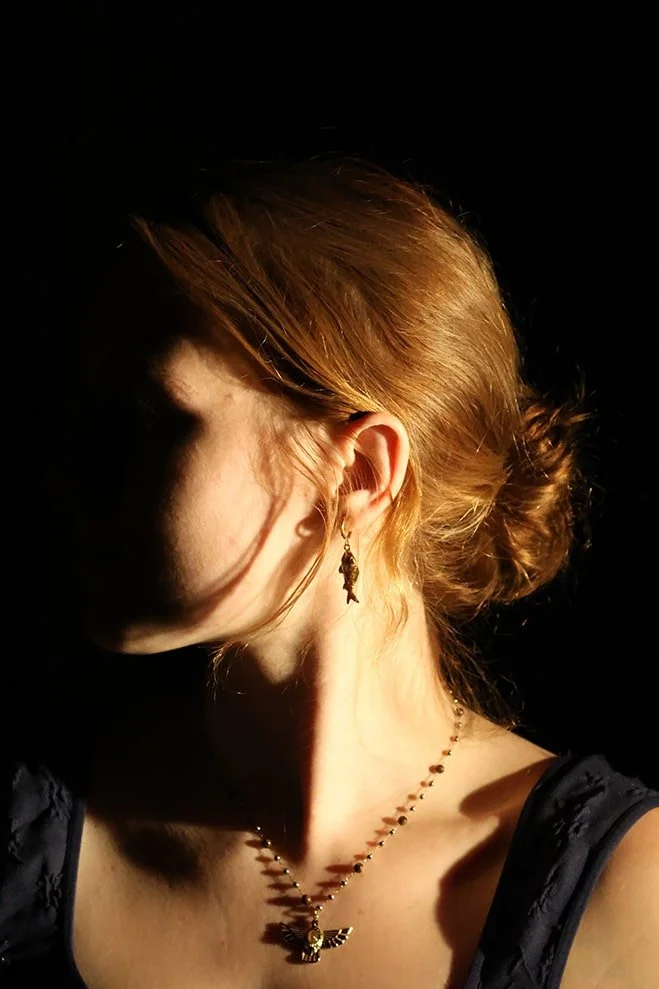 Close-up of a woman with red hair styled in a bun, wearing a necklace and an earring, with her face turned away from the camera and dramatic lighting casting shadows on her neck and shoulder.
