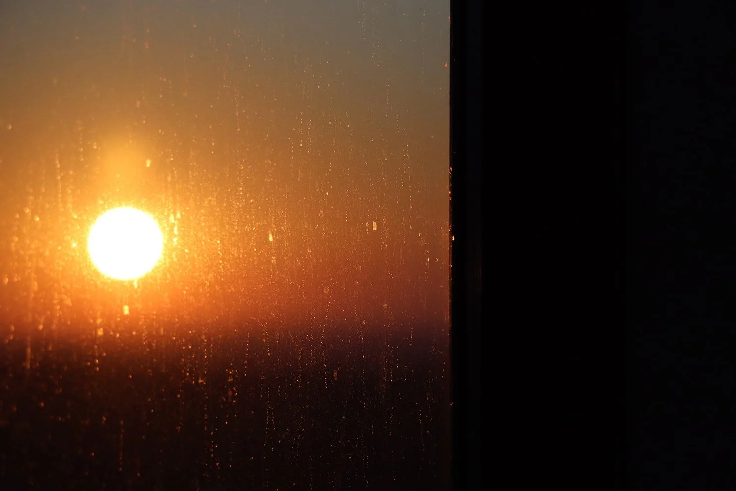 Sunset view through a window with raindrops, showing the bright sun and orange sky.