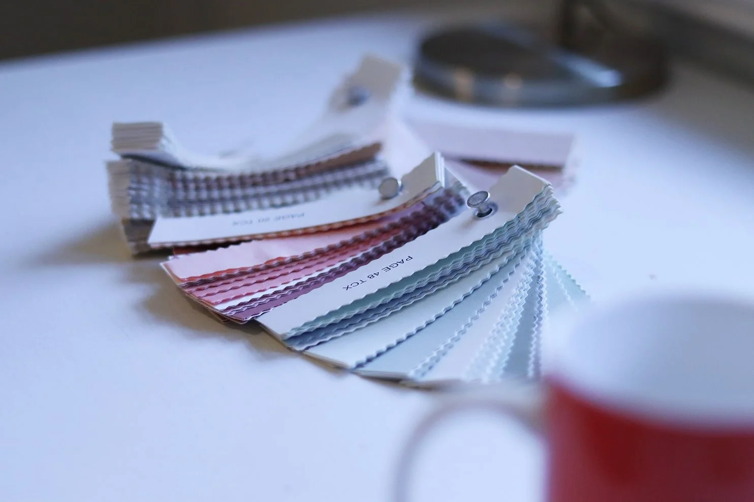 Color swatches of paint sample cards spread out on a white surface with a red cup in the foreground and a paint roller in the background.