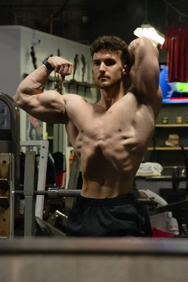 A shirtless man flexing his arm muscles and showing his abs in a gym.