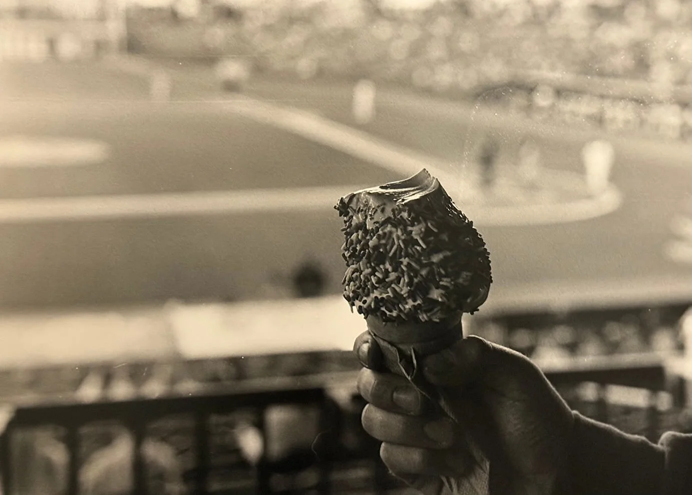 A hand holding an ice cream cone with chocolate sprinkles and a chocolate coating, against a blurred background of what appears to be a dessert shop or street scene.