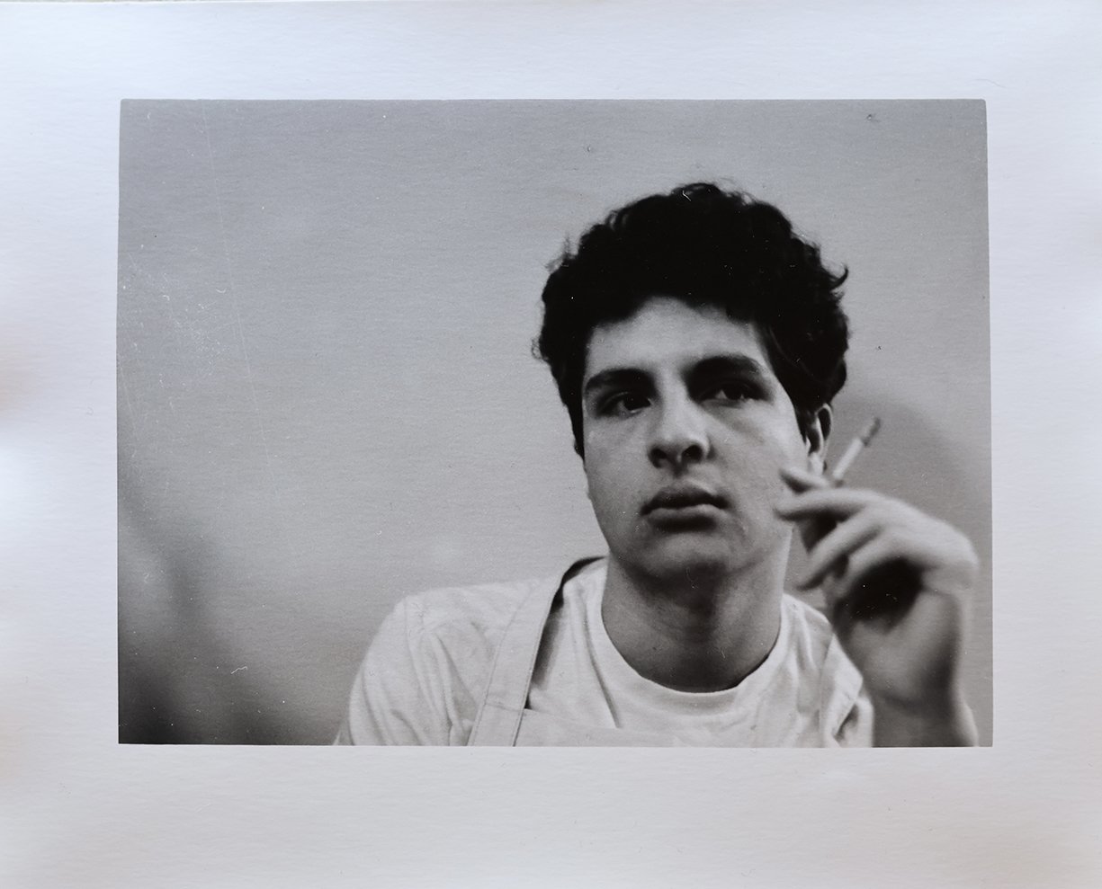 Black and white photograph of a young man with dark, curly hair holding a cigarette near his face, looking directly at the camera with a serious expression, against a plain background.