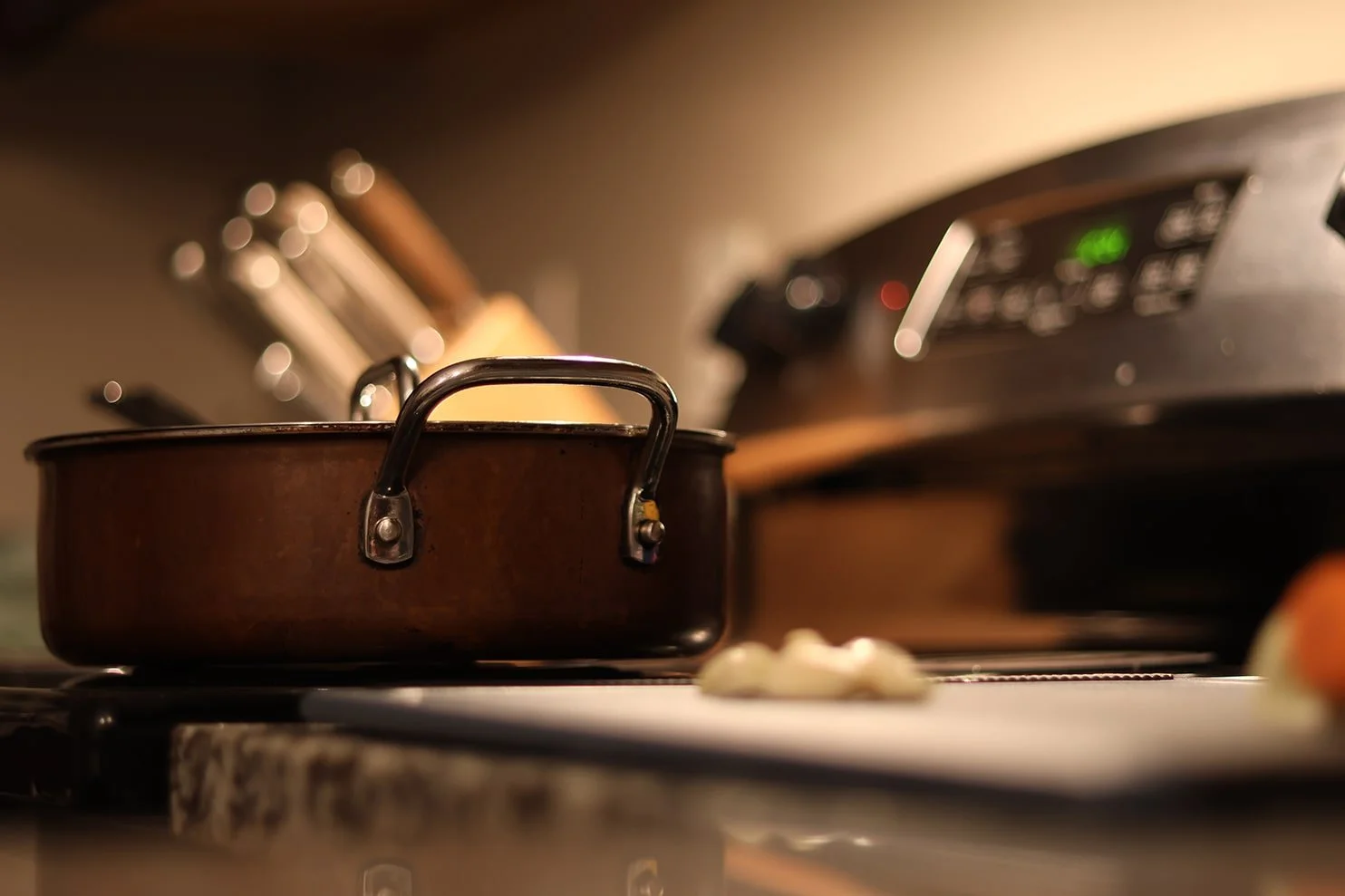 A close-up of a small copper pot on a stovetop with a blurred background of a kitchen, including knives and a toaster.