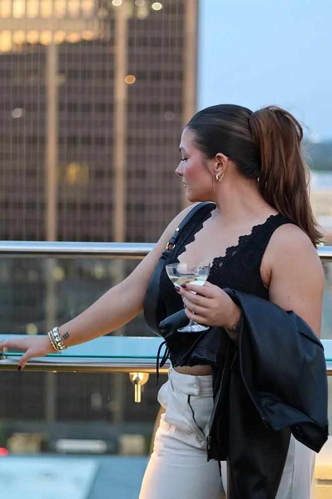 A woman with dark hair tied in a ponytail, wearing a black lace sleeveless top, white pants, and holding a glass of drink, standing on a balcony with a cityscape background during sunset or twilight.