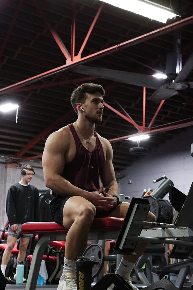A young man with athletic build sitting on a workout bench at the gym, wearing a maroon tank top and black shorts, with his hands clasped. There are gym equipment and another person in the background.