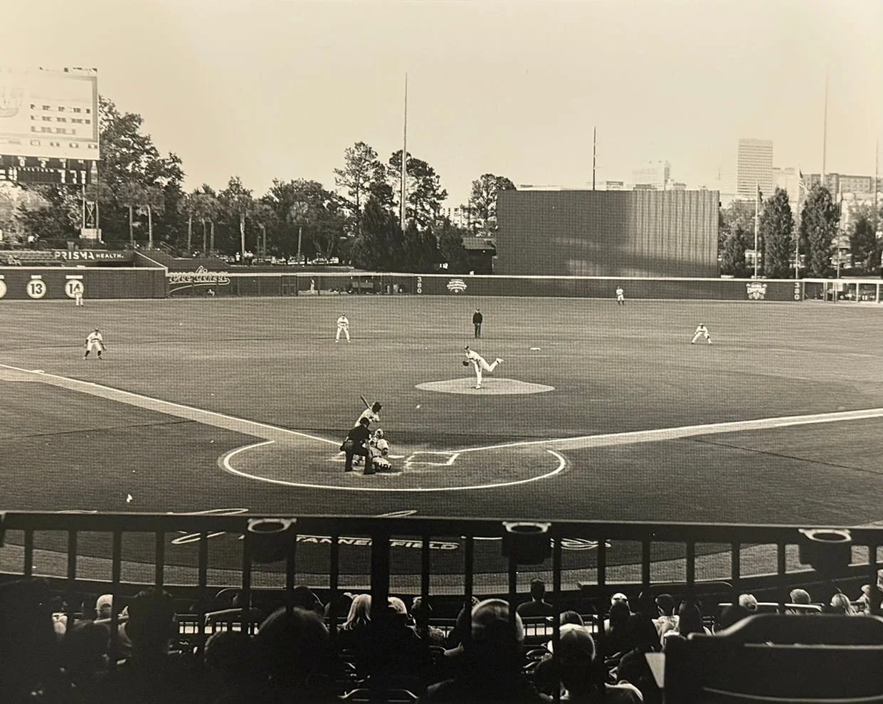 Black and white photo of a baseball game at a stadium, with players on the field, spectators in the stands, and city buildings in the background.