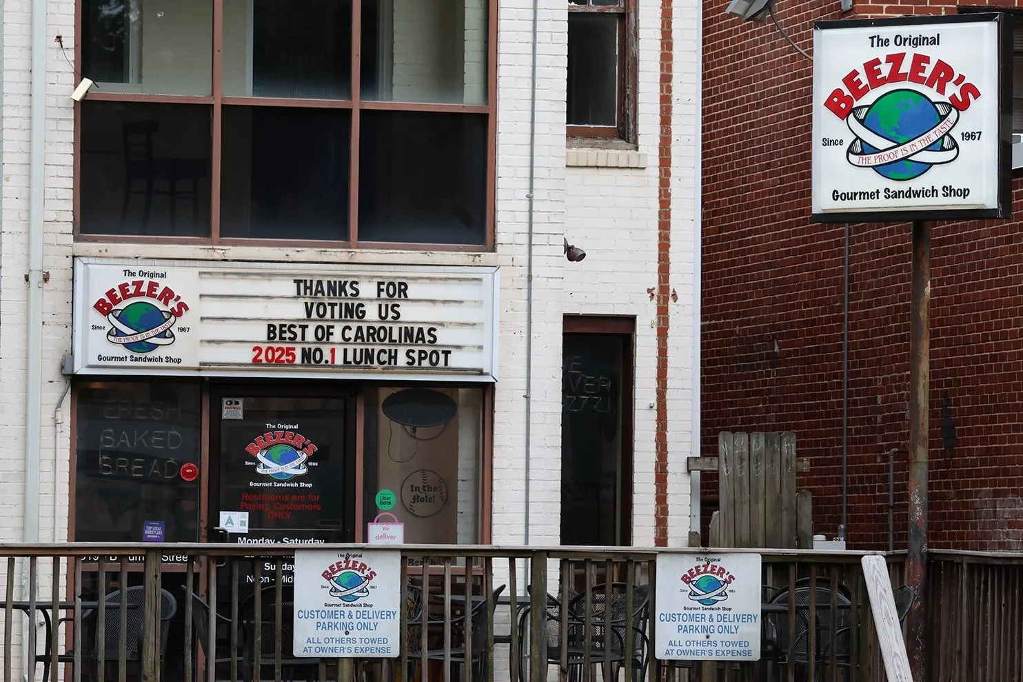 Exterior of Beezer's Gourmet Sandwich Shop with signs, a white brick facade, and a red brick wall. The marquee thanks customers for voting them best in Carolinas and mentions they are the No. 1 lunch spot in 2025. Signs indicate customer and delivery