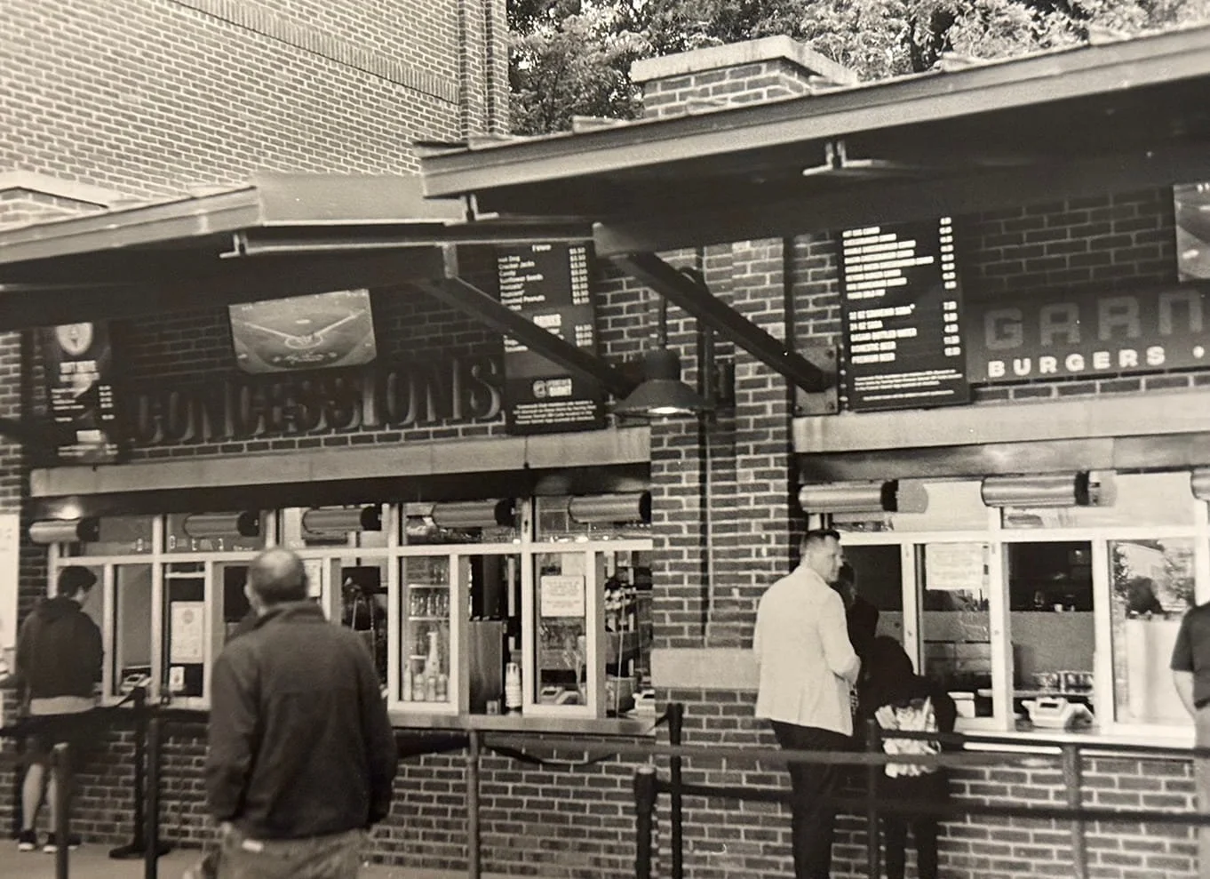 People ordering food at a brick restaurant or fast food stand with menu boards and a visible sign that reads 'BURGERS'.