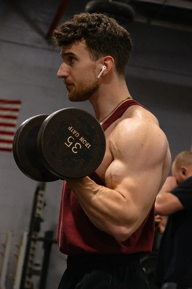 A young man with dark hair and a beard, wearing earbuds, performing a bicep curl with a 35-pound dumbbell in a gym.