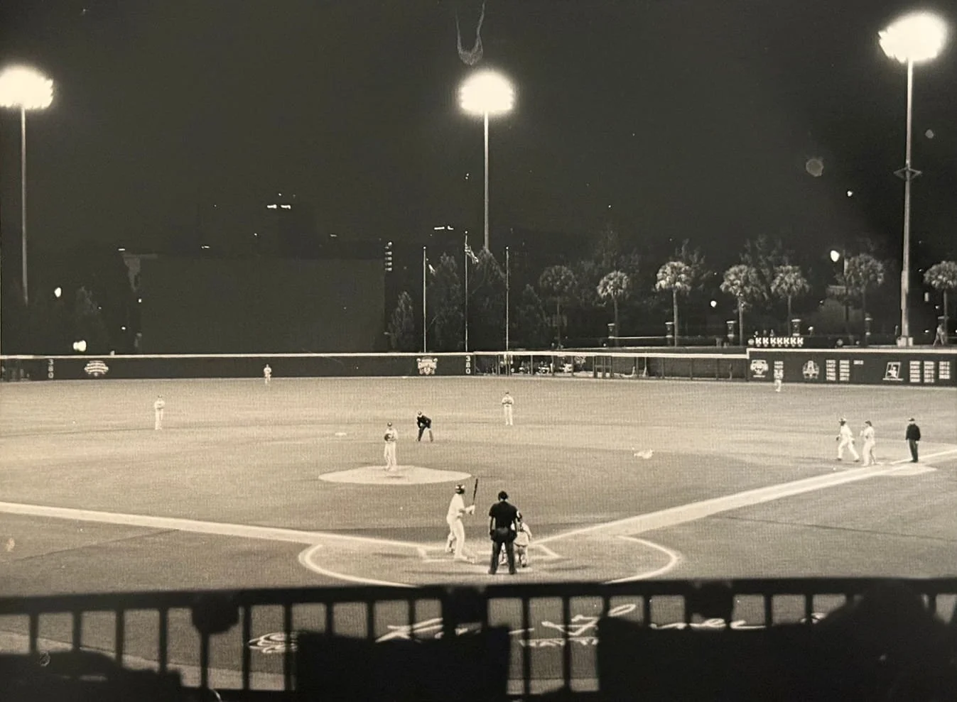 Black and white photo of a baseball game at night, with players on the field and bright stadium lights illuminating the scene.