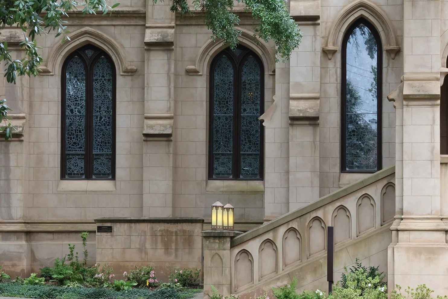 Facade of a stone building with large, gothic-style windows and a staircase, surrounded by greenery and lanterns.