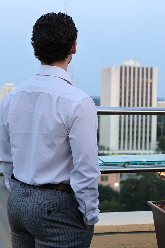 A man with dark hair, wearing a white dress shirt and gray pinstriped trousers, stands on a rooftop or balcony and looks out over a cityscape with tall buildings and a cloudy sky.