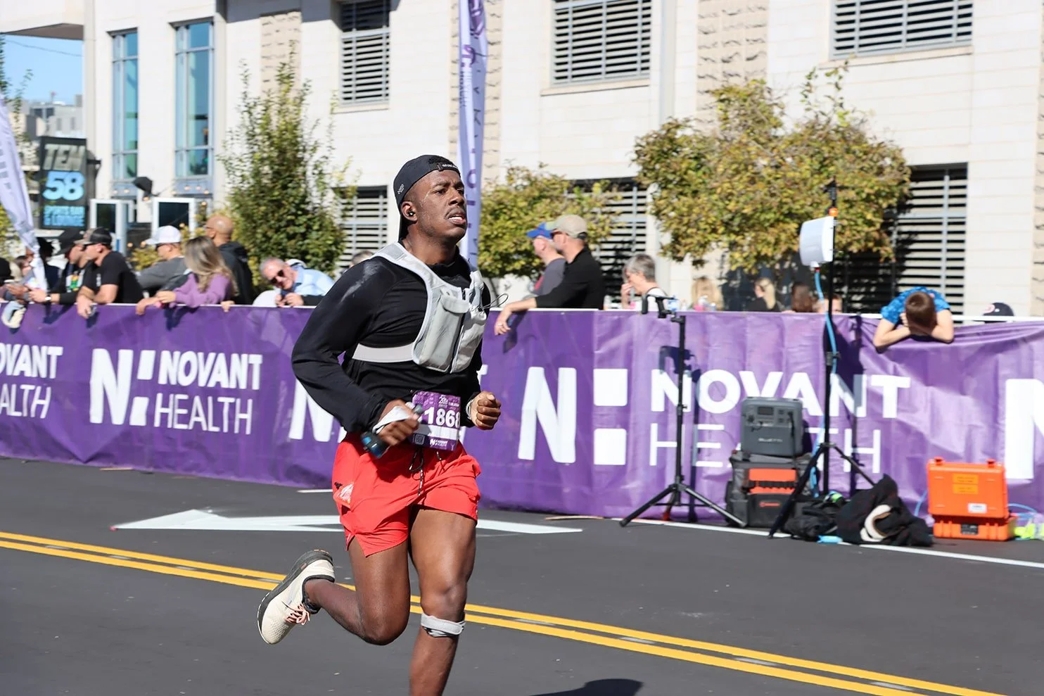 A male runner wearing a black cap, black long sleeve shirt, orange shorts, and a white hydration vest, participating in a marathon, with spectators behind a purple banner that has 'NOVAANT HEALTH' printed on it, along with equipment on the side of th