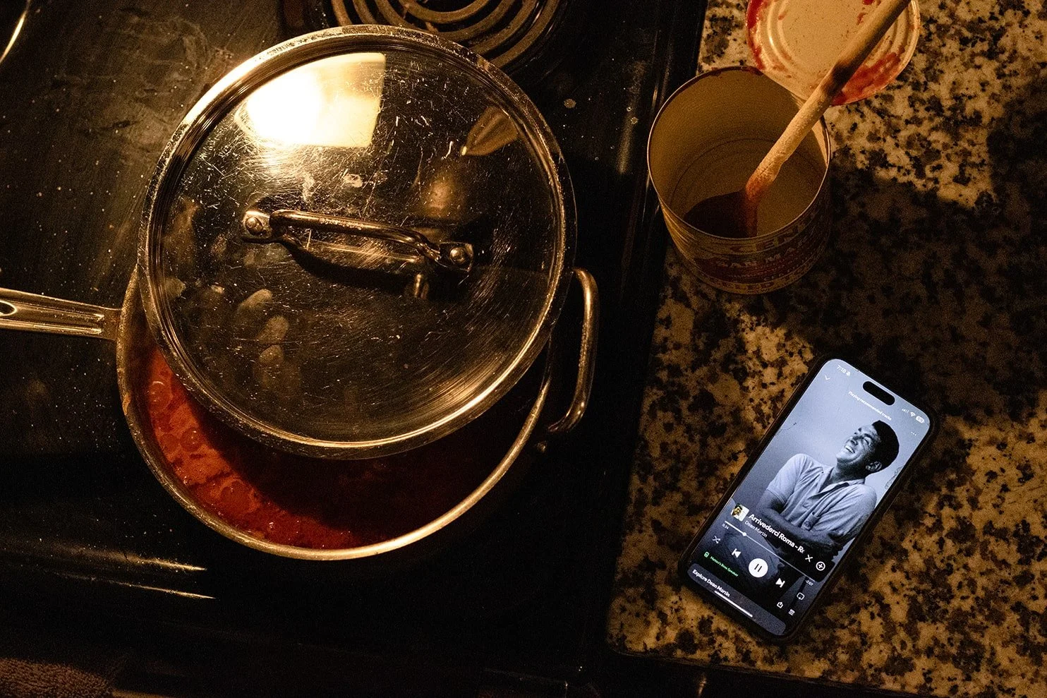 A stove with a pot and a glass with a wooden stirring stick on a speckled granite countertop, with a smartphone displaying a black-and-white image of a smiling man.