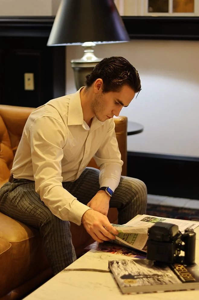 A young man with dark hair, wearing a beige shirt and pattern pants, sitting on a brown sofa, reading a newspaper. There is a black camera and a magazine on the table in front of him, with a lamp and framed pictures in the background.