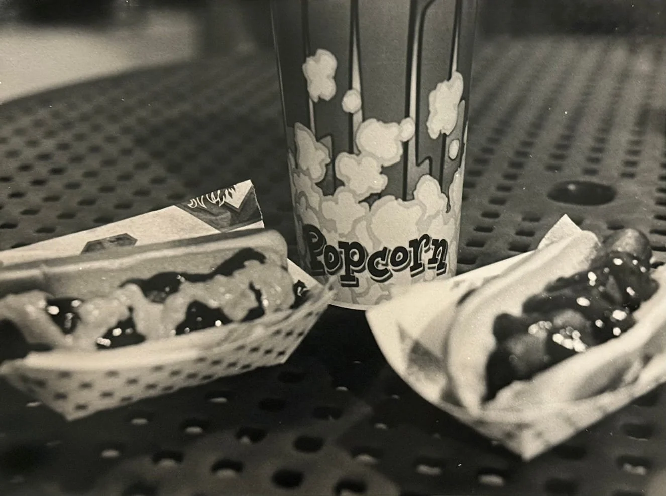 A small popcorn container, two hot dog buns with hot dogs topped with mustard or ketchup, all on a metal perforated table.