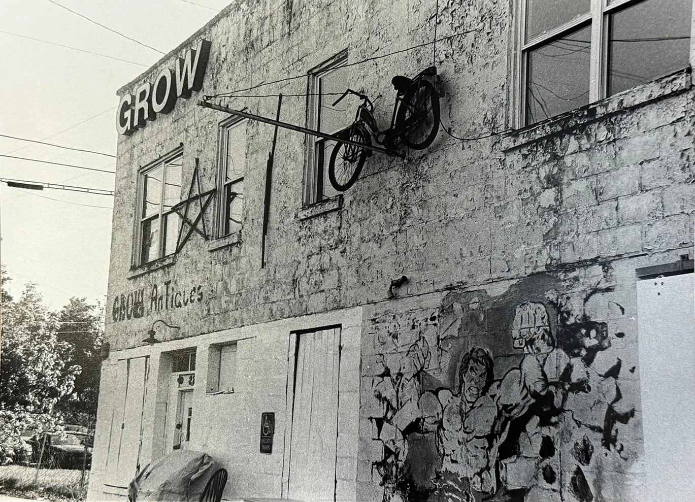 Black and white photo of a building with a sign that reads 'GROW' and a painted mural of people fighting, with a bicycle sculpture hanging on the wall.