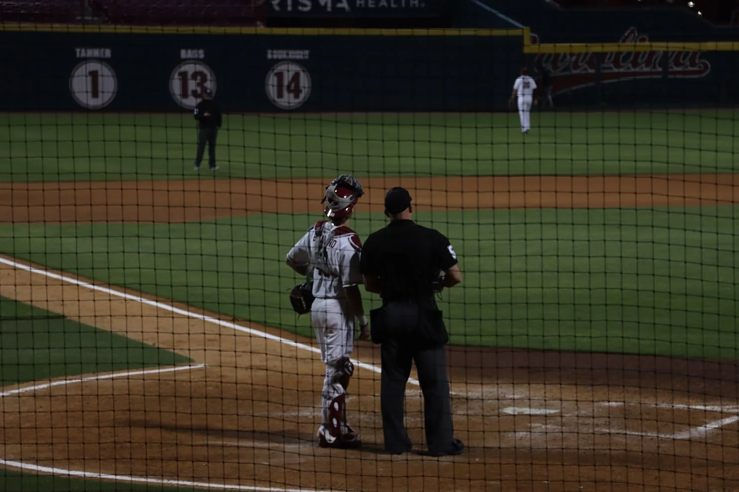 A baseball game at night with a batter, catcher, and umpire near home plate, viewed through a safety net.