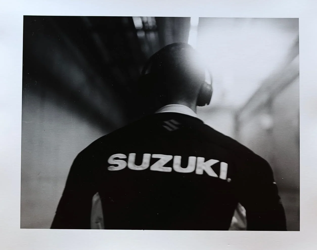 Back view of a person in a Suzuki jacket with overall dark hair, wearing headphones, walking in an indoor environment.