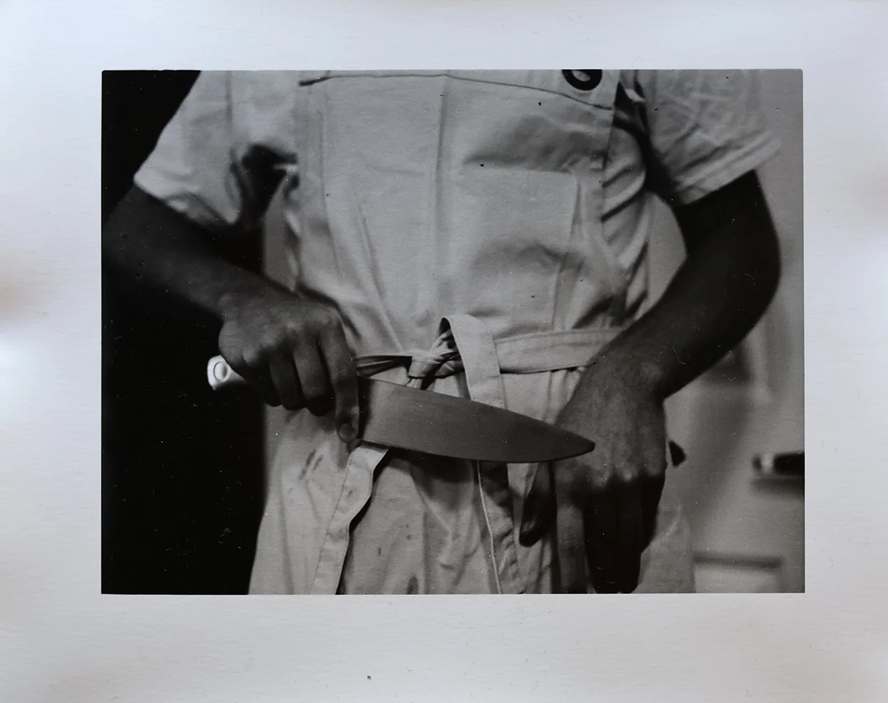 Black and white photo of person wearing a chef's coat and apron, holding a kitchen knife in one hand and a knife sharpener in the other.