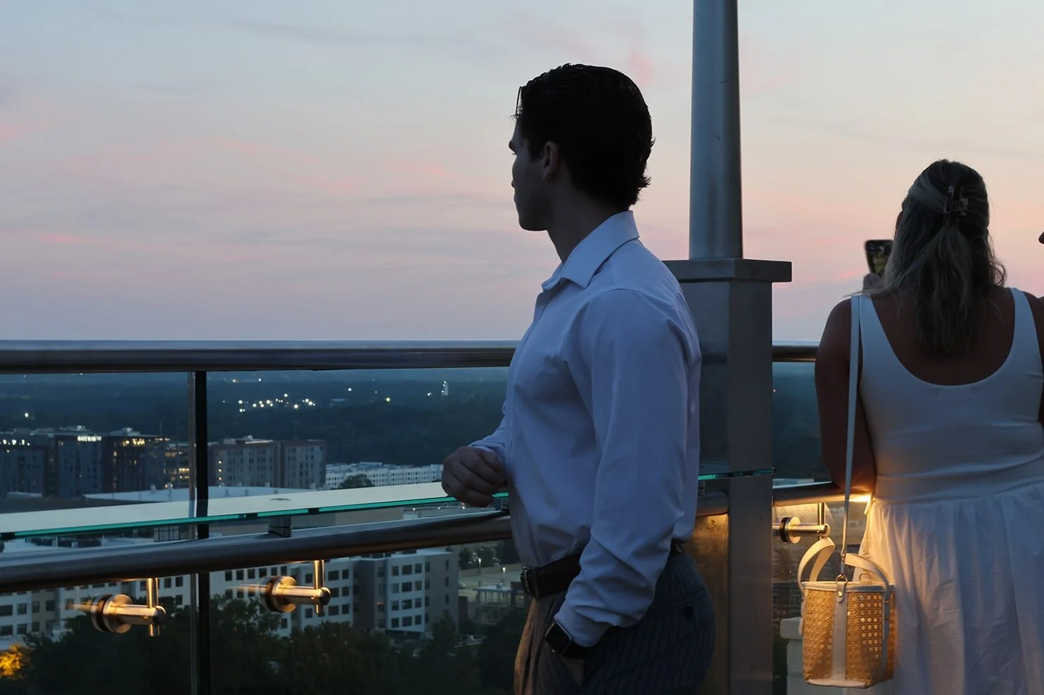 A man in a white shirt and gray pants standing on a rooftop balcony at sunset, looking out over a cityscape, with a woman in a white dress taking a photo or video with her phone.