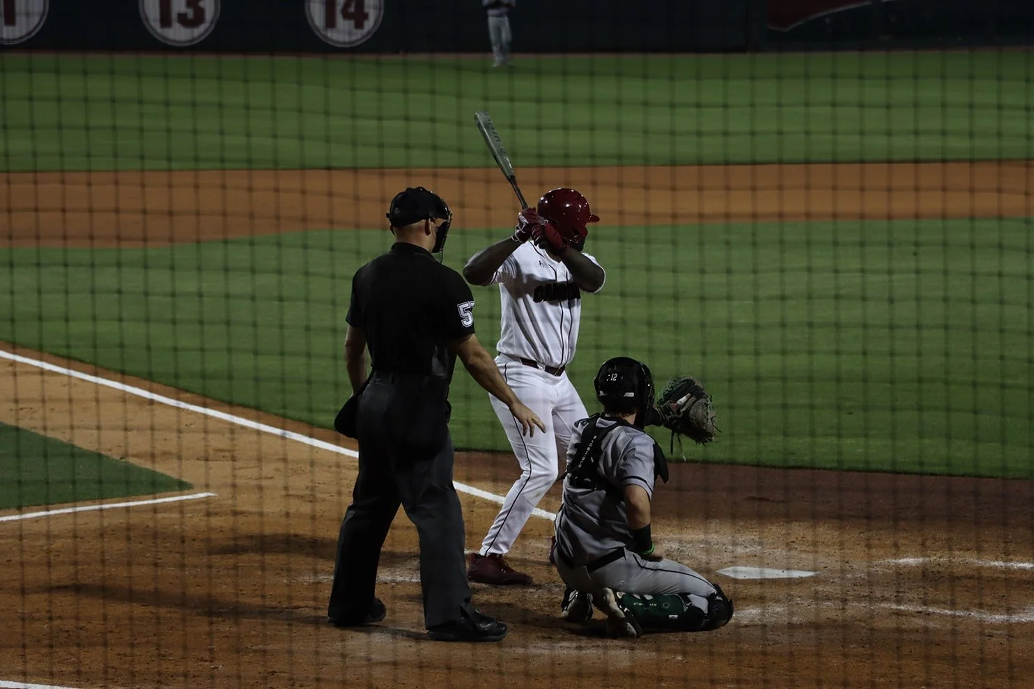 A baseball player stands at home plate preparing to bat during a game, with an umpire and catcher nearby on the field, all wearing uniforms and protective gear.