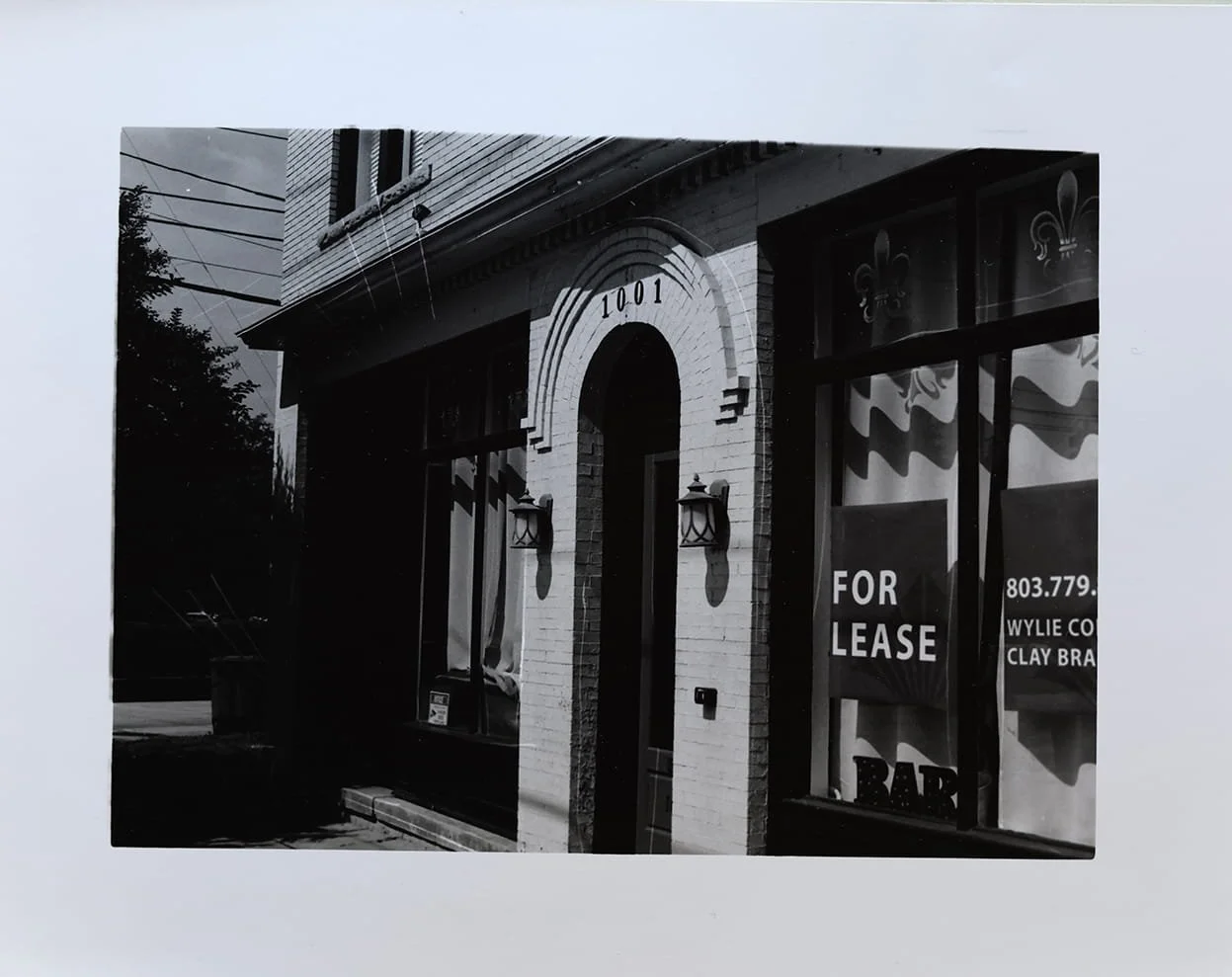 Black and white photo of a commercial building with a sign that says 'FOR LEASE' in the window, and a phone number and company information. The building has an arched entrance with the number 1001 above it, and brick exterior with decorative lanterns