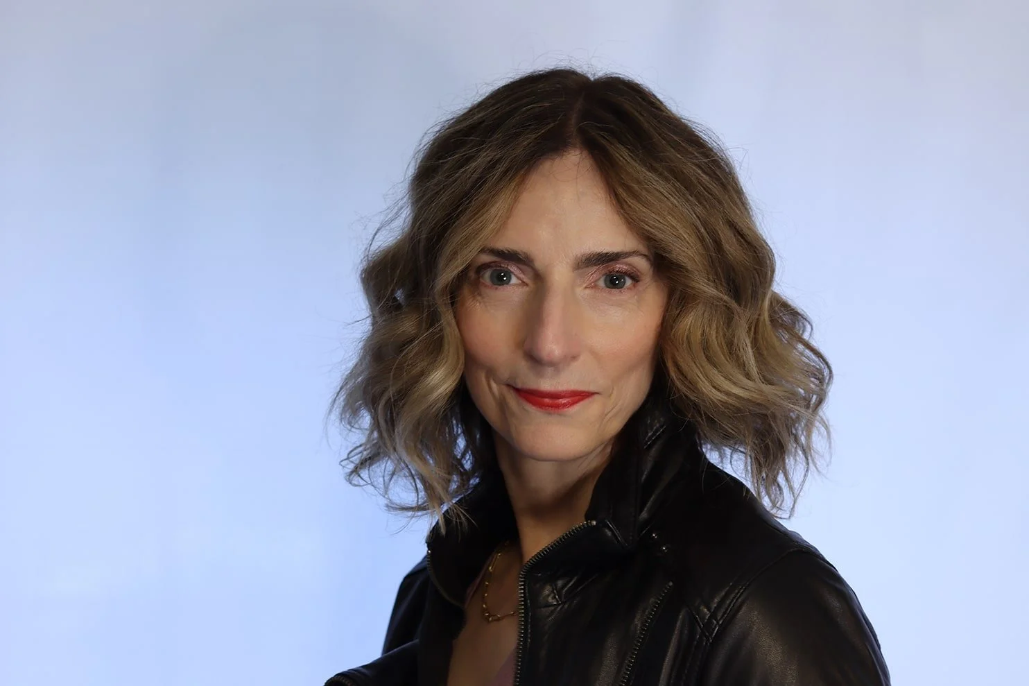 A woman with shoulder-length wavy hair, red lipstick, and a black leather jacket posing against a light blue background.