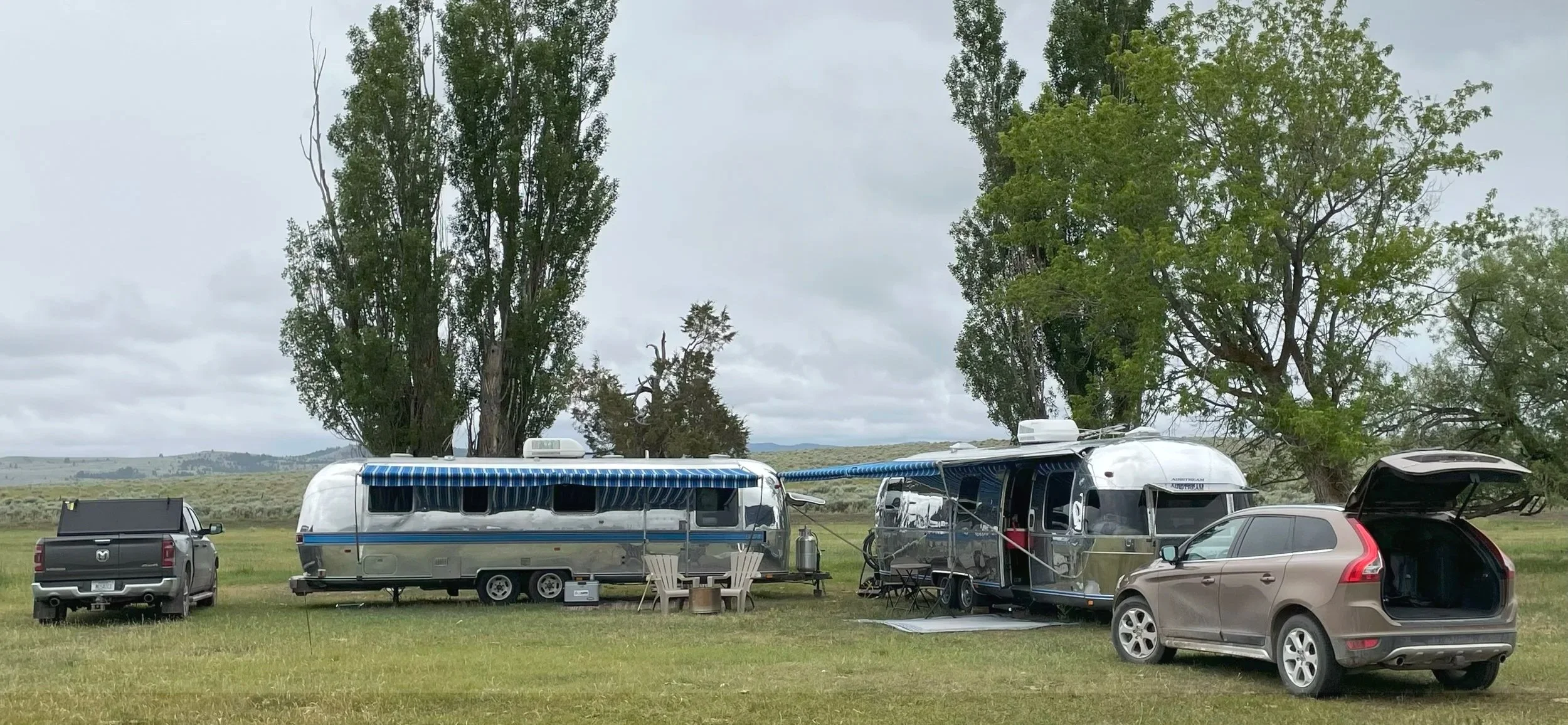 Two remodeled travel trailers pulled by cars, parked on a grassy field with trees in the background on a cloudy day.