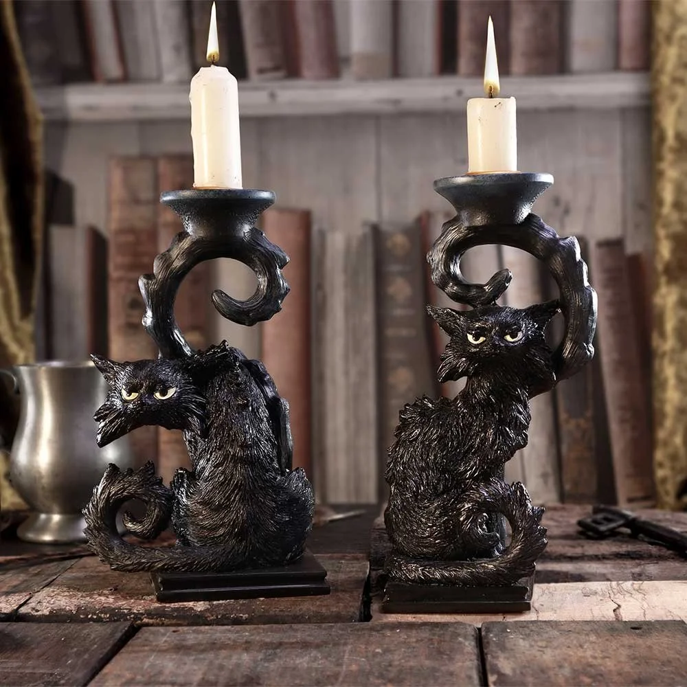 Two black vampire-style candle holders shaped like cats with curled tails and glowing yellow eyes, holding white candles on a rustic wooden table.