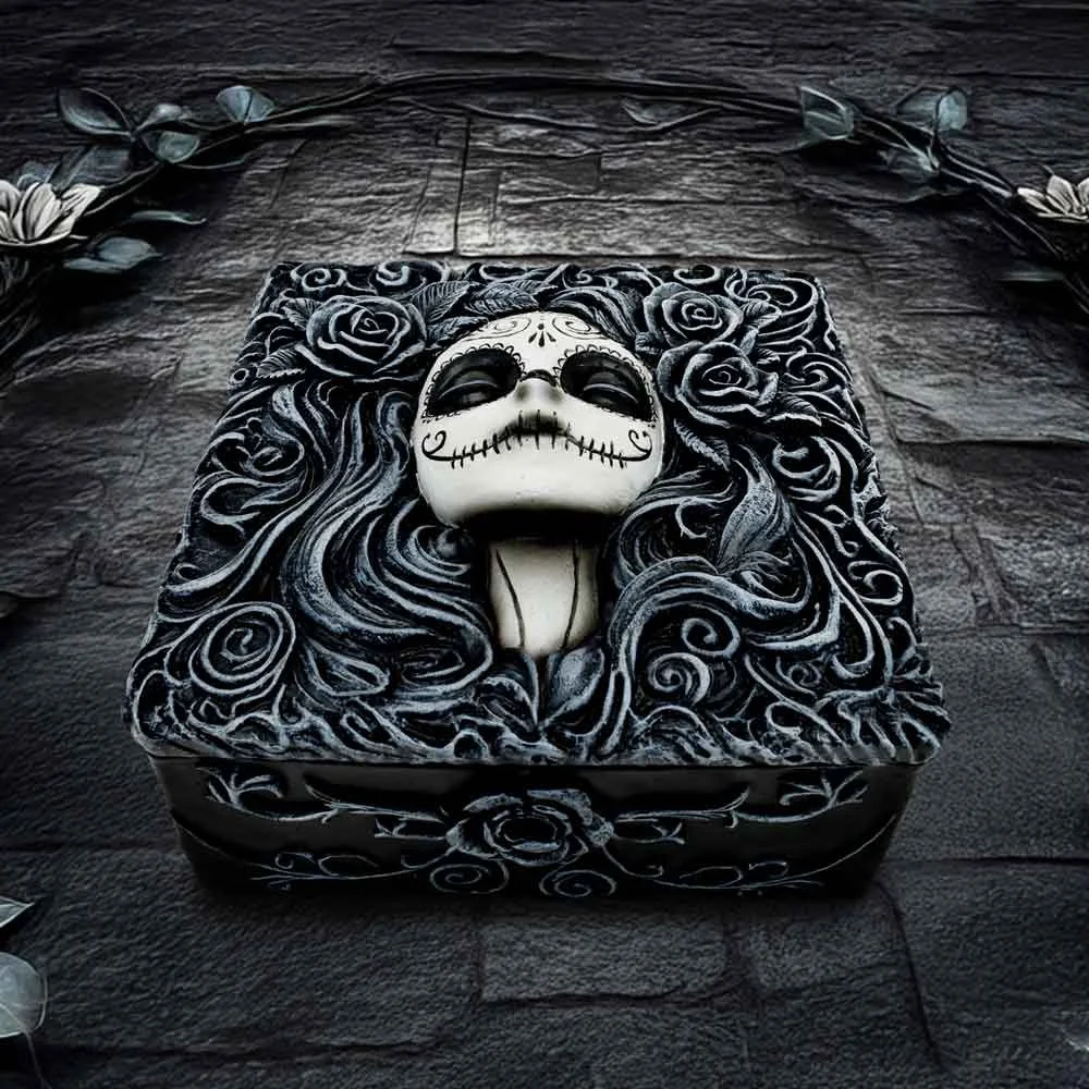 Decorative box with a sugar skull face design, surrounded by detailed roses and swirling patterns, placed on a textured dark stone surface, with a floral border at the top.